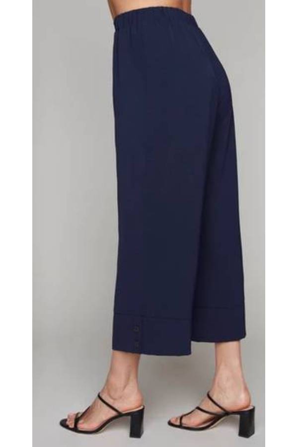 A side waist-down view of a woman wearing the Opal Pant by Compli K in Navy, with a high waist, wide cropped legs, and a button detail on the cuffs. She's standing in front of a grey background.