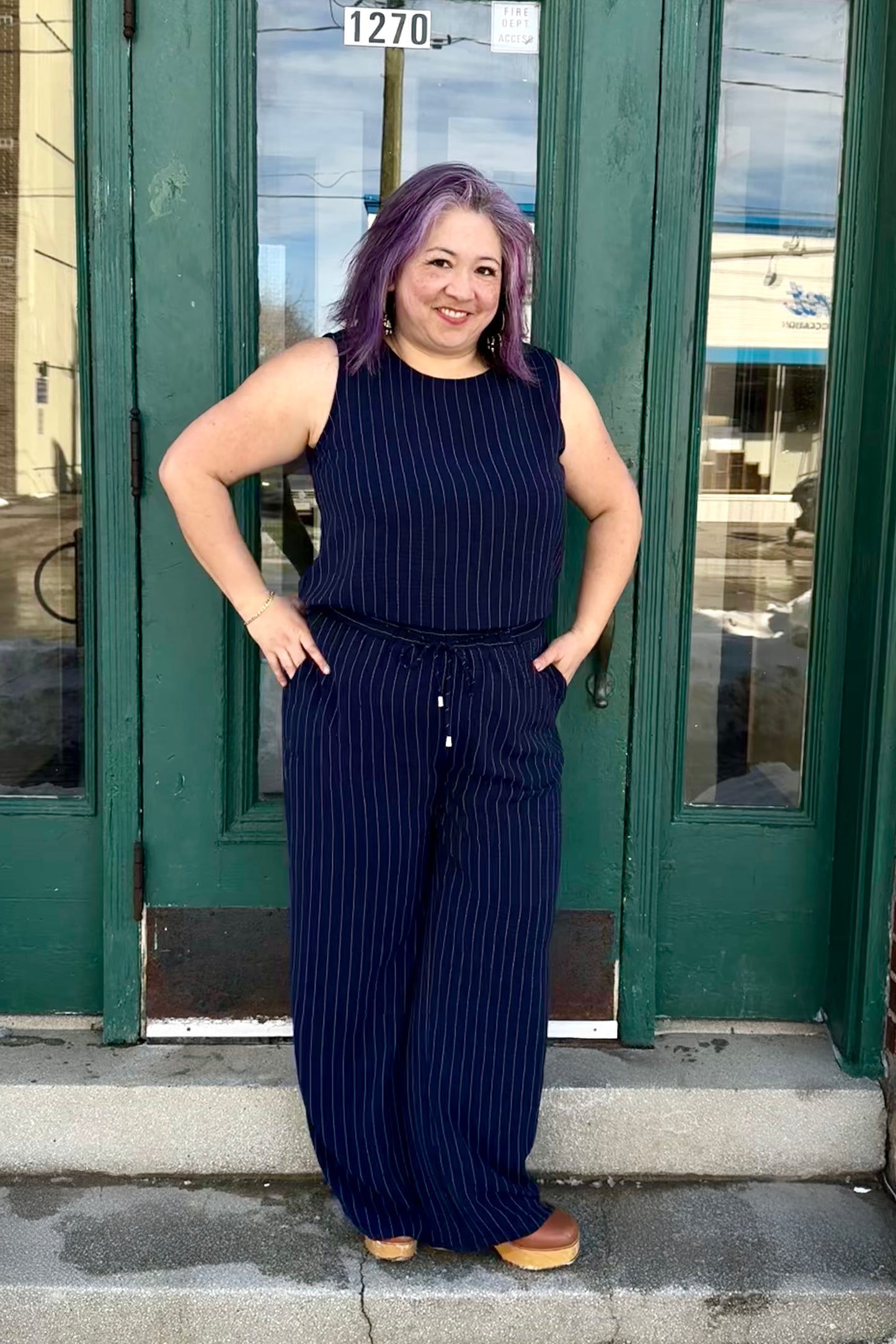 A woman wearing the Ginnie Camisole by Pure Essence, a sleeveless navy/white pinstripe top with a relaxed fit. She is wearing it with the matching
Gabby Pants and standing in front of a green door.