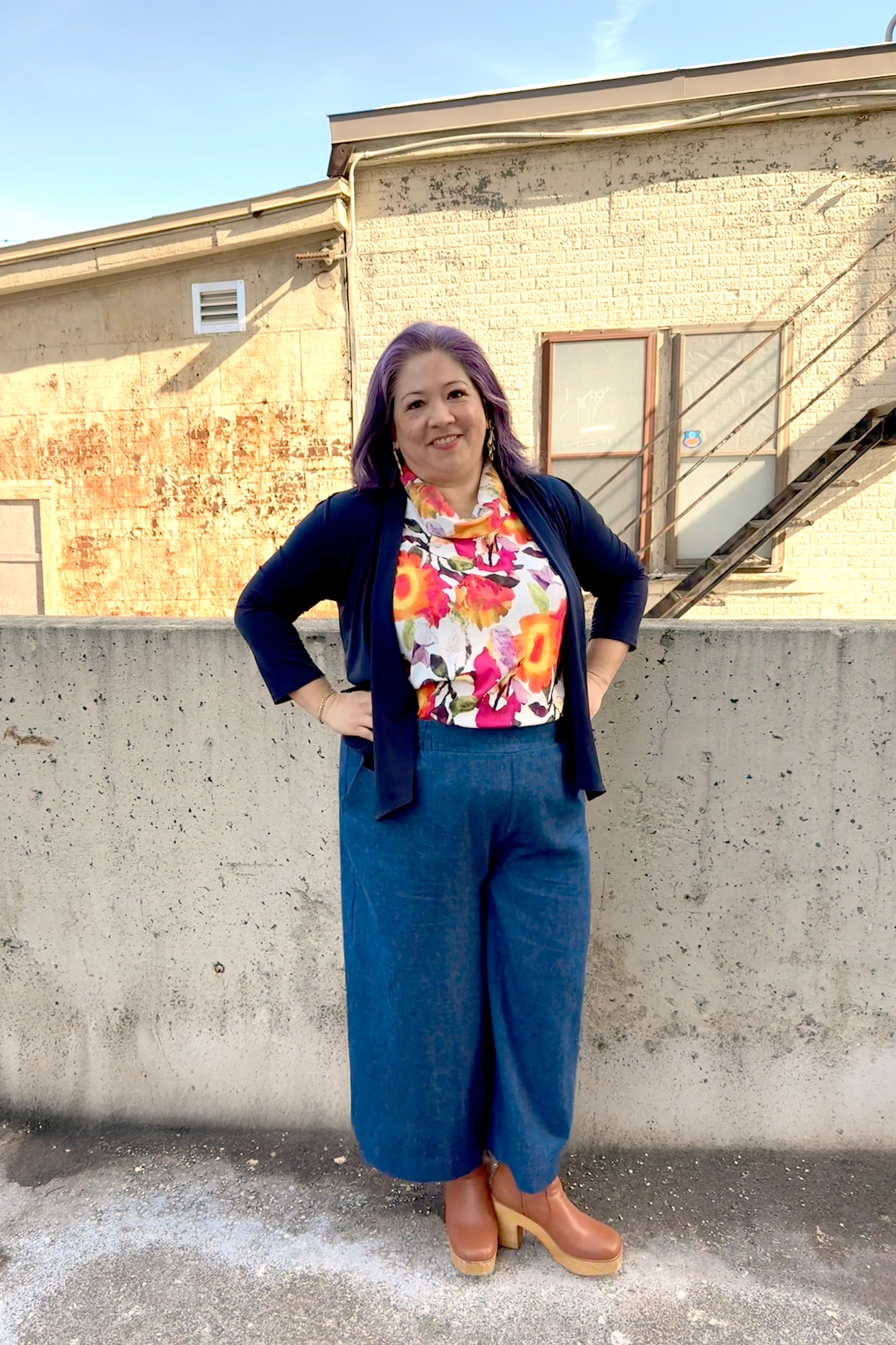 A woman wearing the Gemma top from Pure Essence in Wild Bloom, a floral sleeveless top with a cowl neck. She is wearing it with the Mateo pants from Melow in Denim and a navy cardigan and standing in front of a brick building.