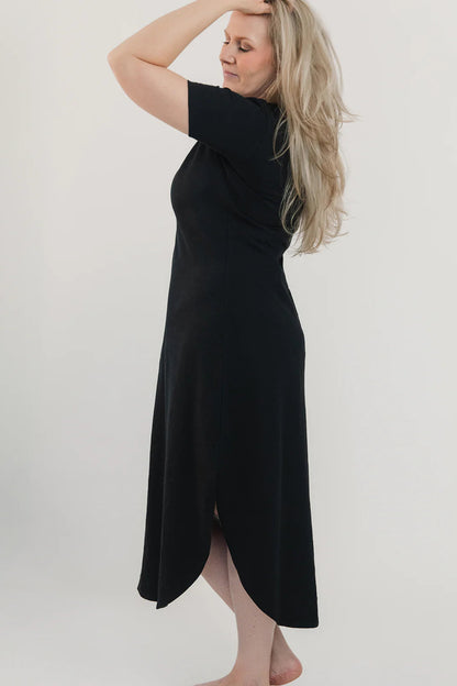 Side view of a woman wearing the Field Dress by Blondie in Black, with short sleeves, a scooped neck, and a contoured hem that hits at mid-calf and cuts up at the sides. She is standing in front of a white background. 