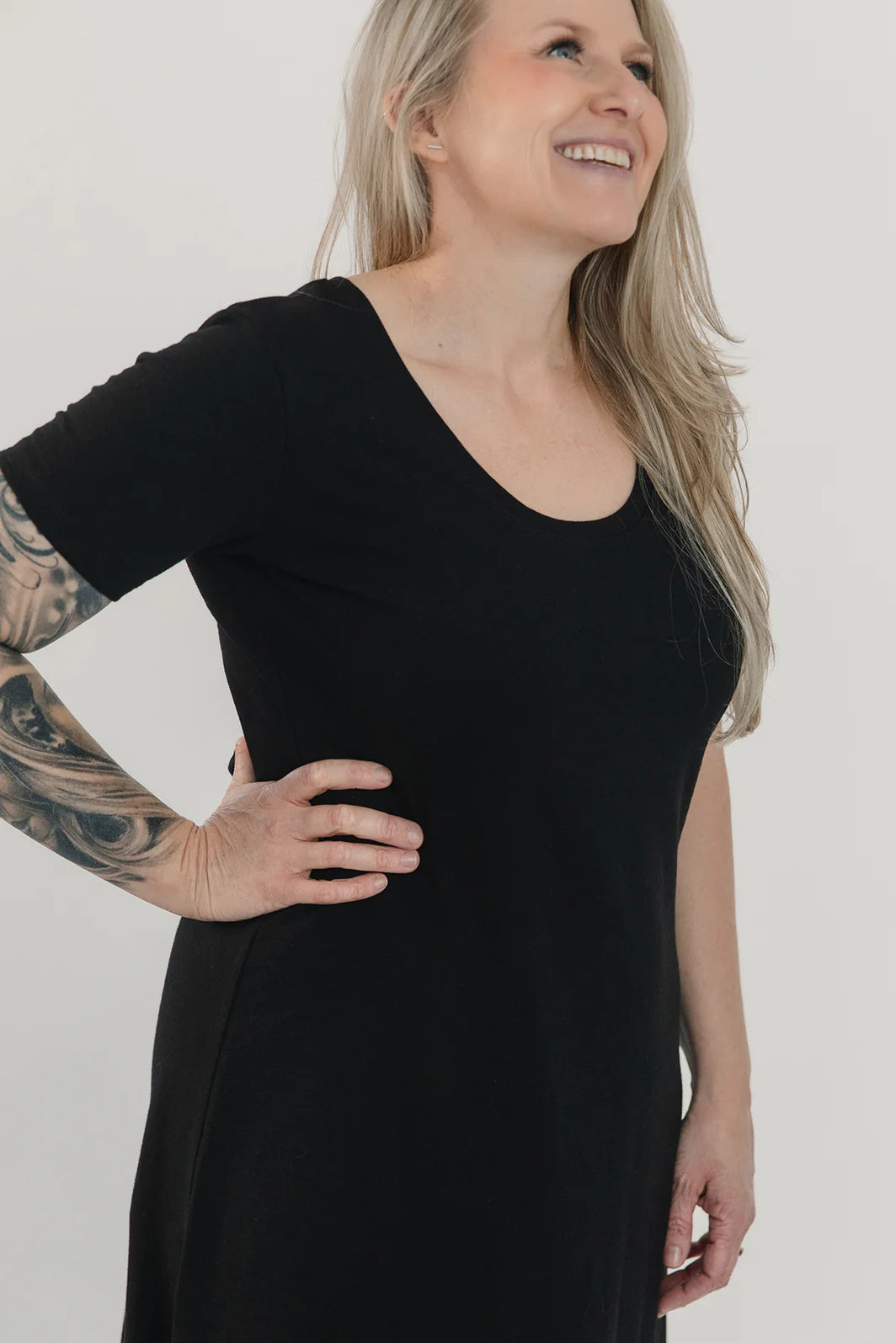 Side close up view of a woman wearing the Field Dress by Blondie in Black, with short sleeves, a scooped neck, and a contoured hem that hits at mid-calf and cuts up at the sides. She is standing in front of a white background. 