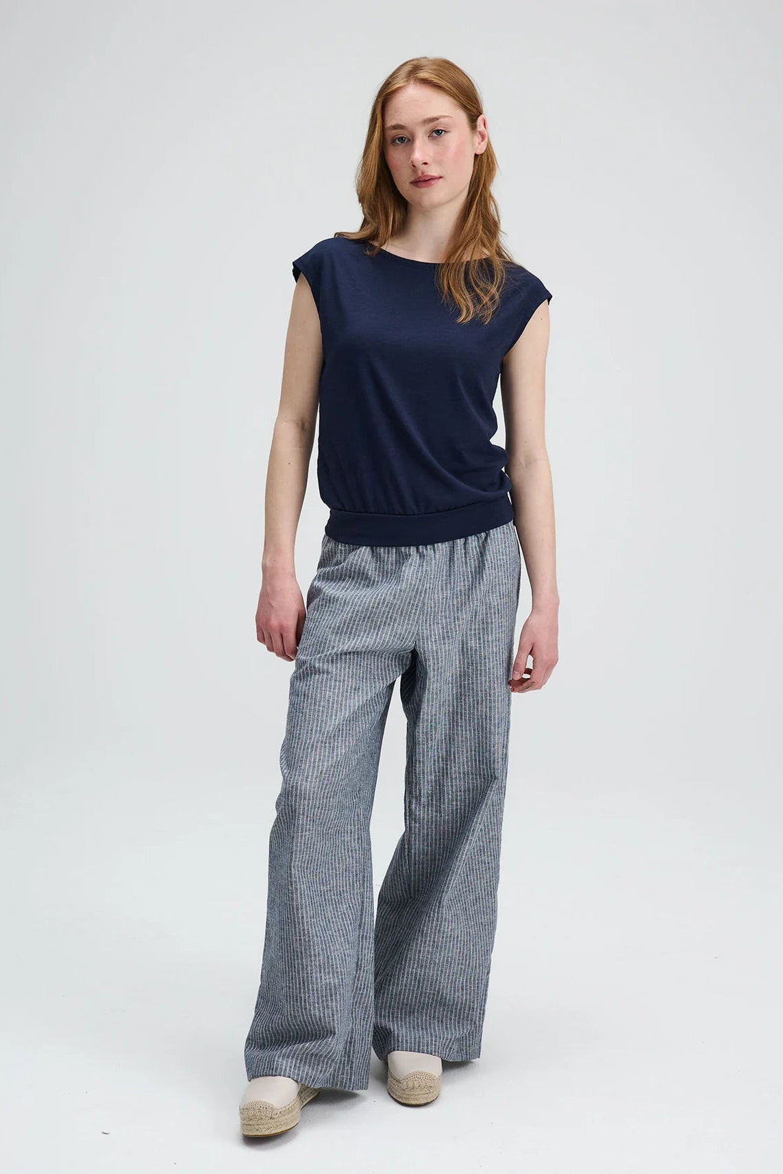 A woman wearing the Liberte pants from Message Factory in Grey stripe, loose fitting wide legged pants with an elastic waist and side pockets. She is wearing them with a navy shirt and standing against a white background
