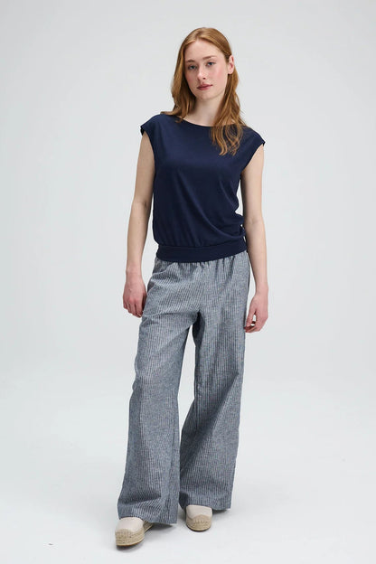 A woman wearing the Batiscan Top by Message Factory in Navy, with short dropped sleeves and a wide neckline, plus a band at the hem. She is wearing it with the Liberte pants in Grey Stripe and standing against a light grey background  