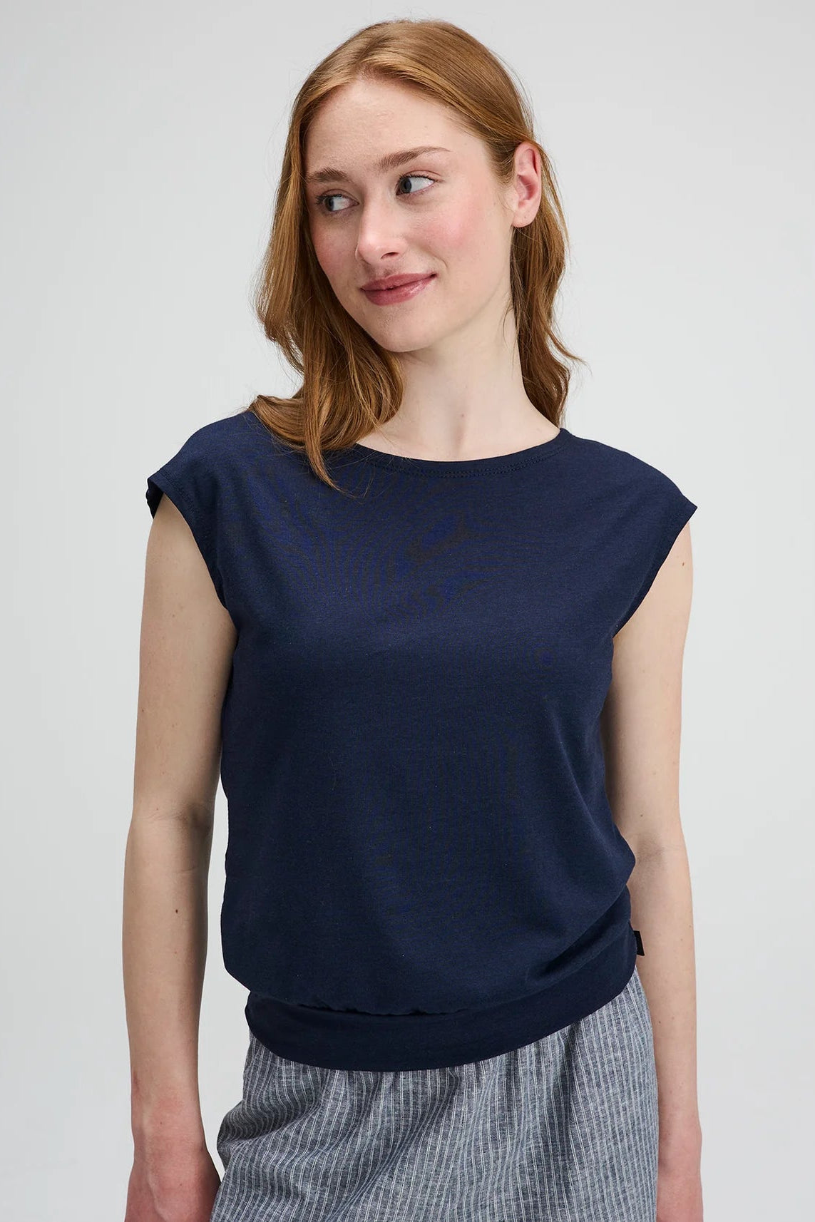 Waist up view of a woman wearing the Batiscan Top by Message Factory in Navy, with short dropped sleeves and a wide neckline, plus a band at the hem. She is wearing it with the Liberte pants in Grey Stripe and standing against a light grey background  