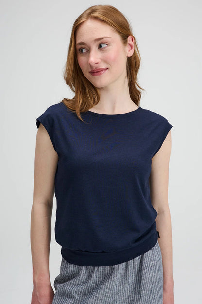 Waist up view of a woman wearing the Batiscan Top by Message Factory in Navy, with short dropped sleeves and a wide neckline, plus a band at the hem. She is wearing it with the Liberte pants in Grey Stripe and standing against a light grey background  