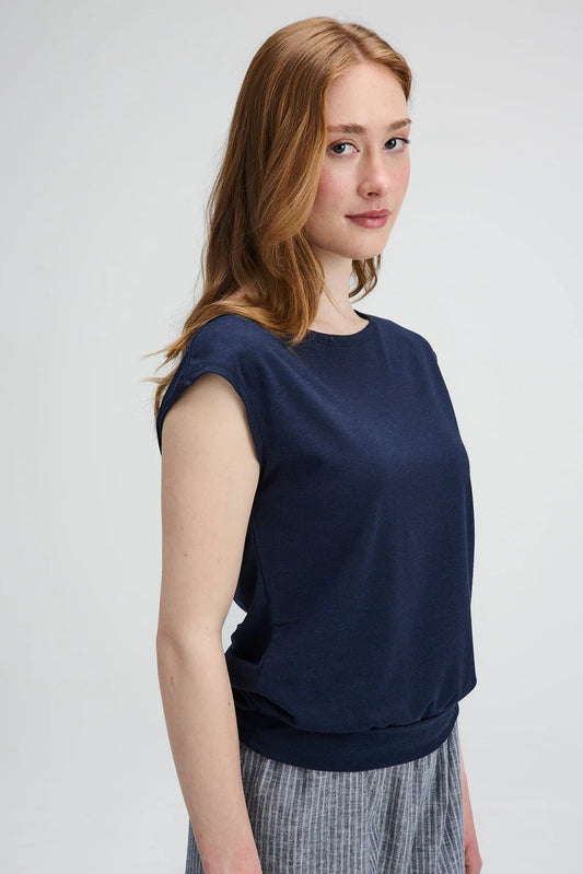 Waist up view of a woman wearing the Batiscan Top by Message Factory in Navy, with short dropped sleeves and a wide neckline, plus a band at the hem. She is wearing it with the Liberte pants in Grey Stripe and standing against a light grey background  