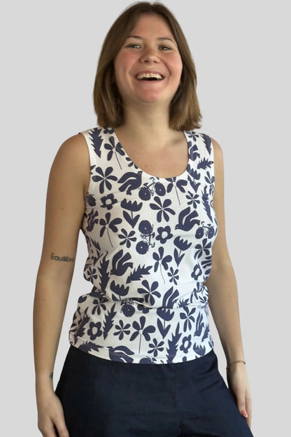 A woman wearing the Mimosa Tank Top by Message Factory, a sleeveless top in a blue floral print. She is wearing it with Navy pants and standing in front of a plain background.
