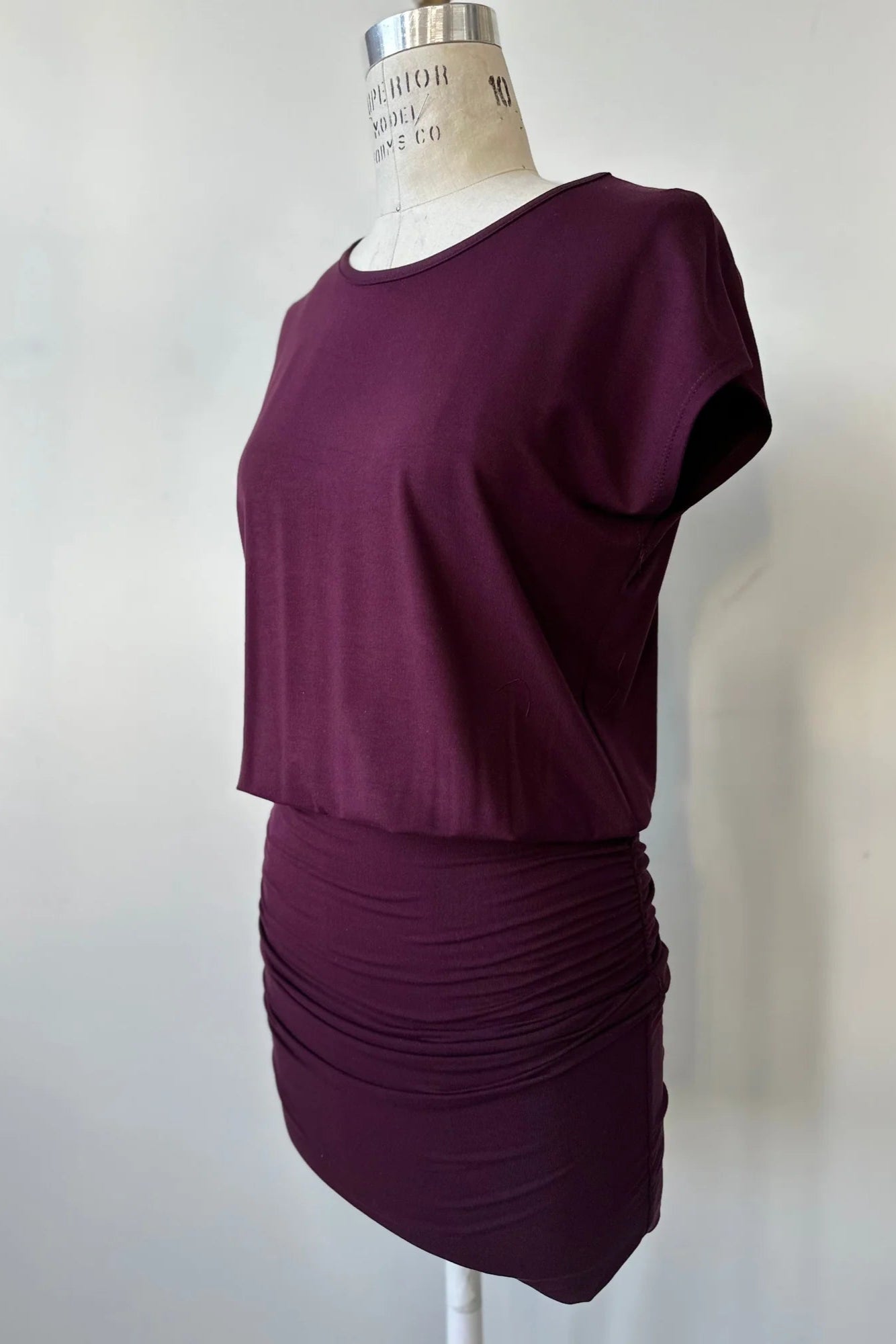 The Gabriel Tunic by Cherry Bobin in Burgundy is shown on a mannequin in front of a white background. It features gathers at the lower sides for an adjustable length, a wide neck, and short extended sleeves. 