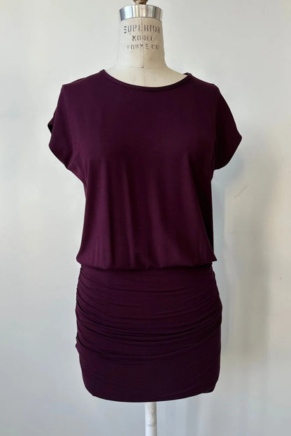 The Gabriel Tunic by Cherry Bobin in Burgundy is shown on a mannequin in front of a white background. It features gathers at the lower sides for an adjustable length, a wide neck, and short extended sleeves. 