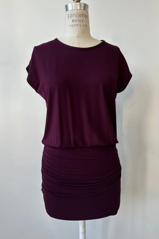 The Gabriel Tunic by Cherry Bobin in Burgundy is shown on a mannequin in front of a white background. It features gathers at the lower sides for an adjustable length, a wide neck, and short extended sleeves. 