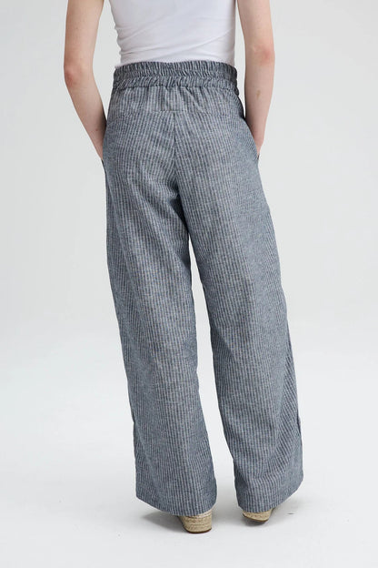 Back waist down view of A woman wearing the Liberte pants from Message Factory in Grey stripe, loose fitting wide legged pants with an elastic waist and side pockets. She is wearing them with a white shirt and standing against a white background