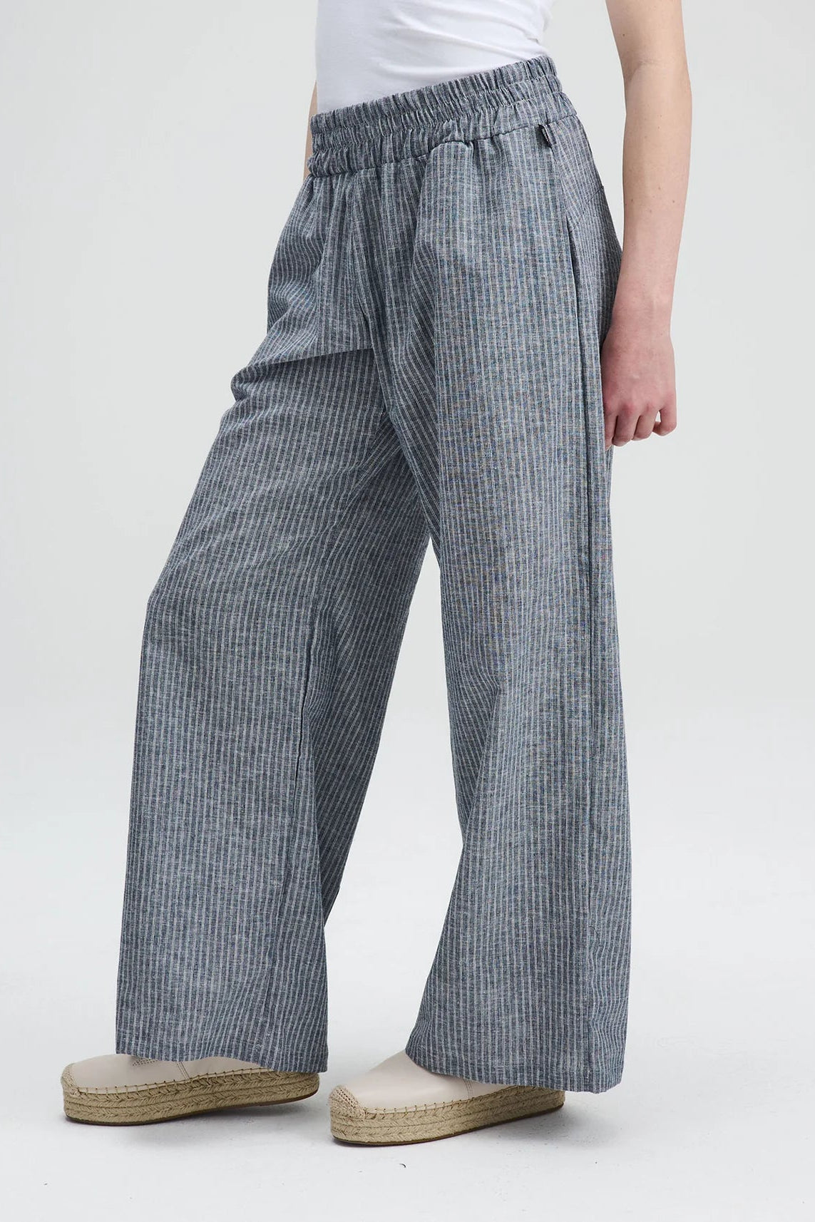 Waist down view of A woman wearing the Liberte pants from Message Factory in Grey stripe, loose fitting wide legged pants with an elastic waist and side pockets. She is wearing them with a white shirt and standing against a white background