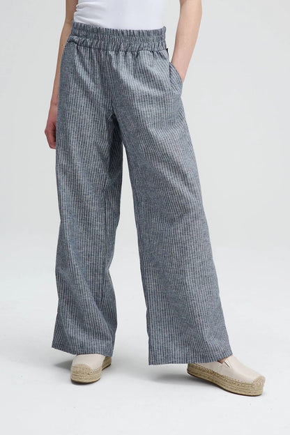 Waist down view of A woman wearing the Liberte pants from Message Factory in Grey stripe, loose fitting wide legged pants with an elastic waist and side pockets. She is wearing them with a white shirt and standing against a white background