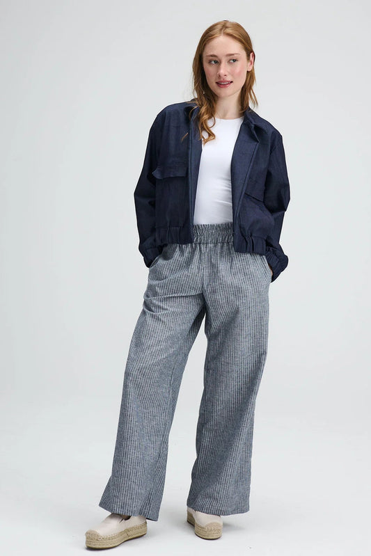 A woman wearing the Liberte pants from Message Factory in Grey stripe, loose fitting wide legged pants with an elastic waist and side pockets. She is wearing them with a navy jacket and a white shirt and standing against a white background