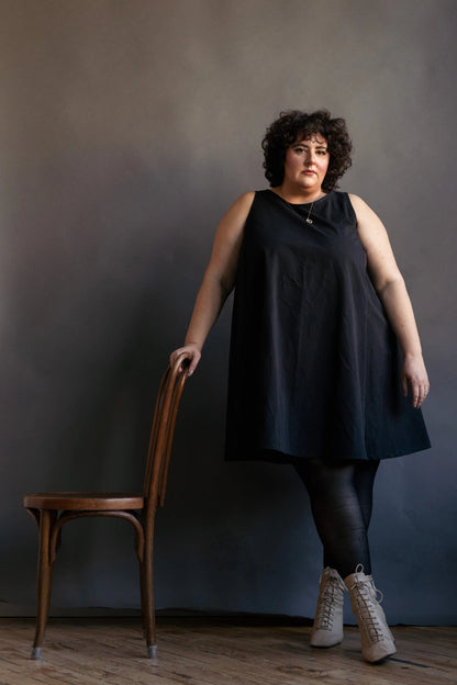 A woman wearing the Caro Dress by Lights of All in Black, a sleeveless knee-length trapeze dress with a round neck at the front and a V at the back. She is wearing it with black leggings and standing with her arm resting on a chair in front of a dark background. 