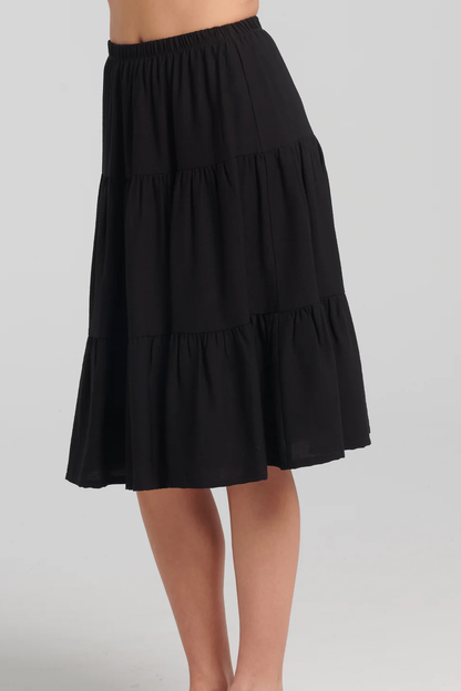 Waist down side view of a woman wearing the Al-Uzza Skirt by Kollontai in Black,  a tiered skirt with an elastic waist and a below-the-knee length, standing against a white background 
