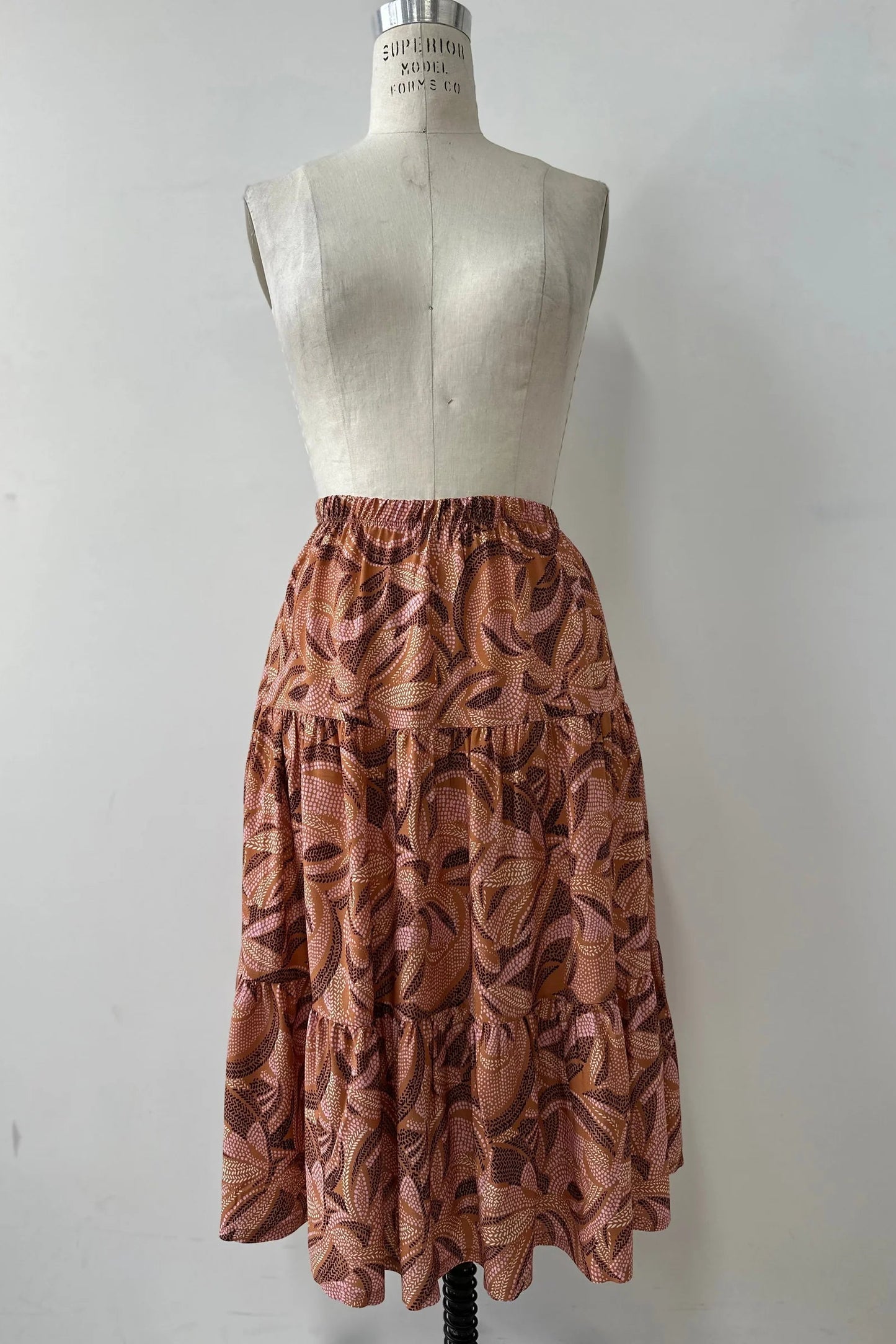 The Al-Uzza Skirt by Kollontai in Cinnamon print, a tiered skirt with an elastic waist and a below-the-knee length, is shown on a mannequin in front of a white background. 