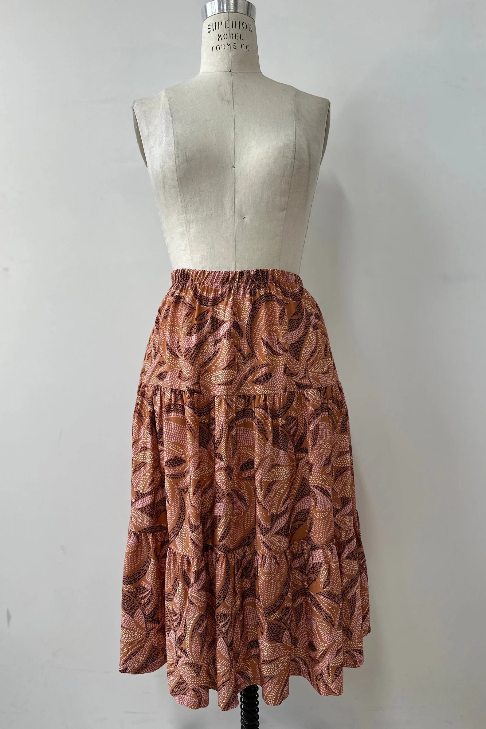 The Al-Uzza Skirt by Kollontai in Cinnamon print, a tiered skirt with an elastic waist and a below-the-knee length, is shown on a mannequin in front of a white background. 