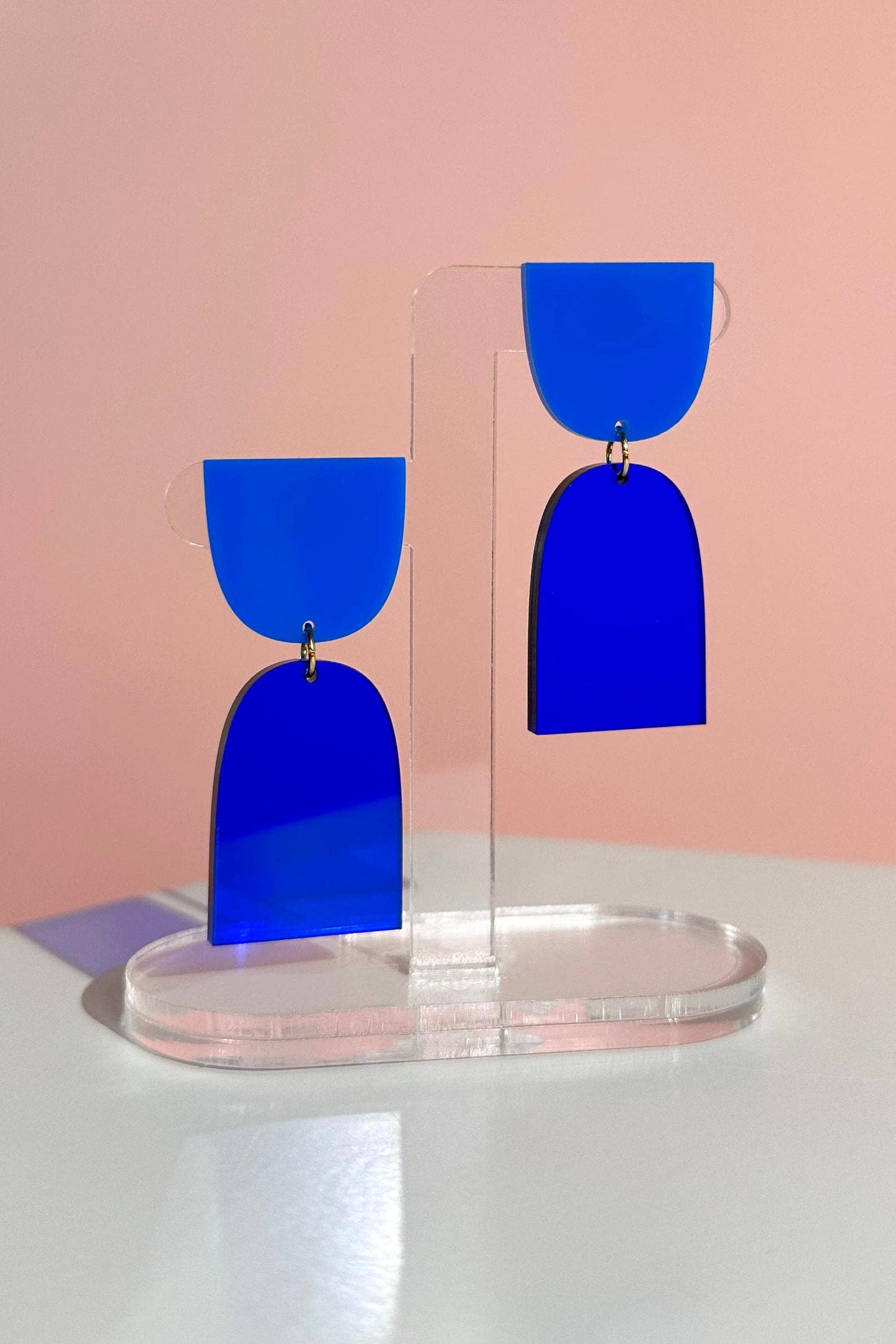 Blue earrings on a clear stand against a pink background