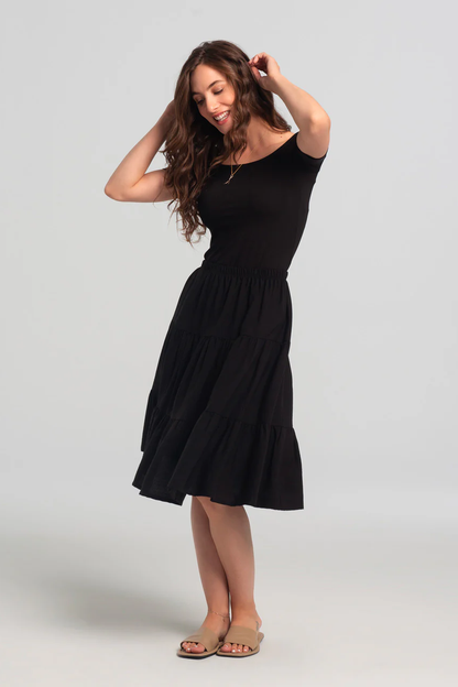 A woman wearing the Al-Uzza Skirt by Kollontai in Black,  a tiered skirt with an elastic waist and a below-the-knee length, with a black top, standing in front of a white background 