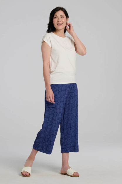 A woman wearing the Eowyn T-Shirt by Kollontai in Off White with blue capri pants standing against a grey background. 