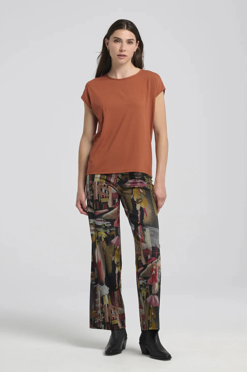 A woman wearing the Eowyn T-Shirt by Kollontai in Rust with patterned pants standing against a grey background. 