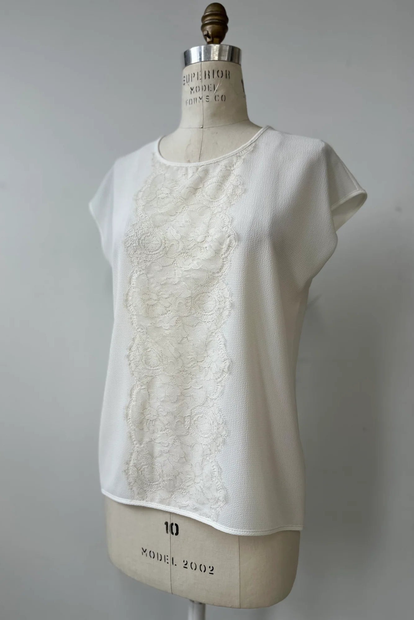 The Pearl T-shirt by Kollontai in White, with short extended sleeves, a relaxed fit, and a lace applique down the front, is show on a mannequin in front of a white background. 