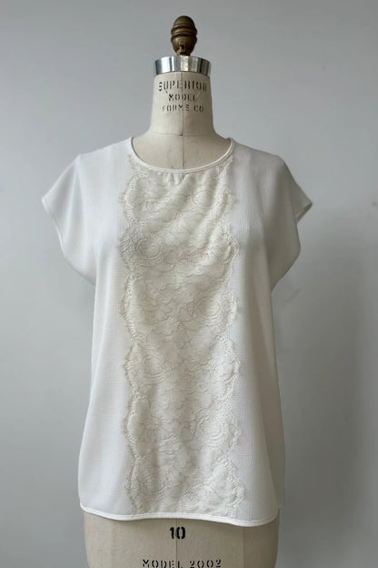 The Pearl T-shirt by Kollontai in White, with short extended sleeves, a relaxed fit, and a lace applique down the front, is show on a mannequin in front of a white background. 