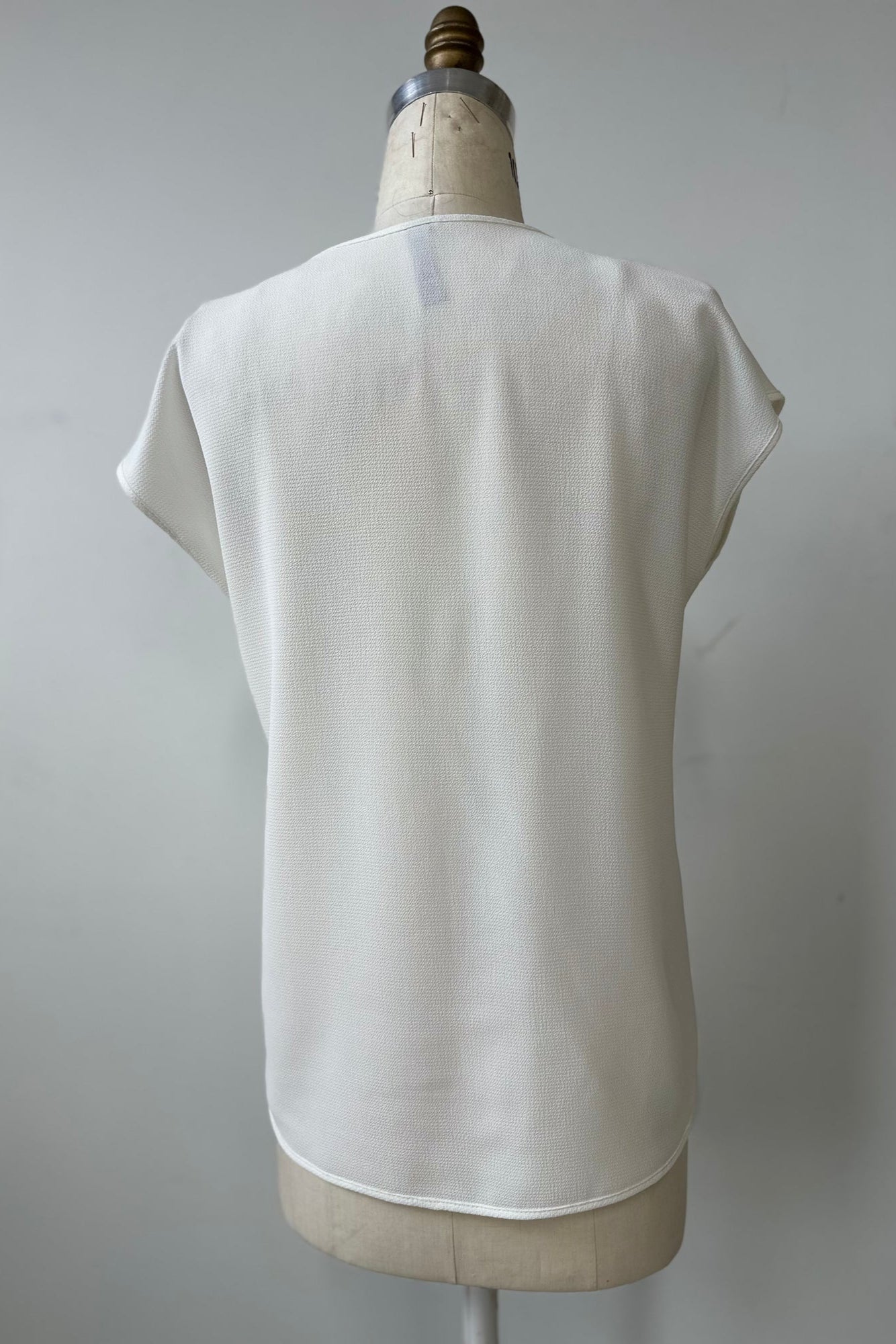 A back view of the Pearl T-shirt by Kollontai in White, with short extended sleeves, a relaxed fit, and a lace applique down the front, is show on a mannequin in front of a white background. 