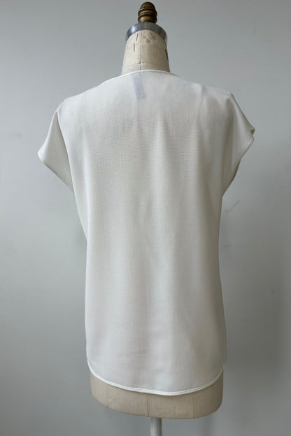 A back view of the Pearl T-shirt by Kollontai in White, with short extended sleeves, a relaxed fit, and a lace applique down the front, is show on a mannequin in front of a white background. 