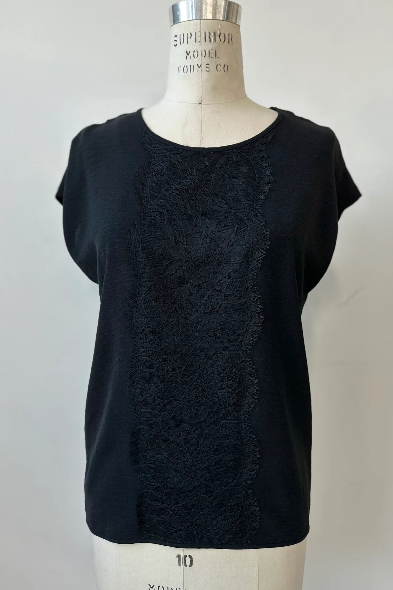 The Pearl T-shirt by Kollontai in Black, with short extended sleeves, a relaxed fit, and a lace applique down the front, is show on a mannequin in front of a white background. 