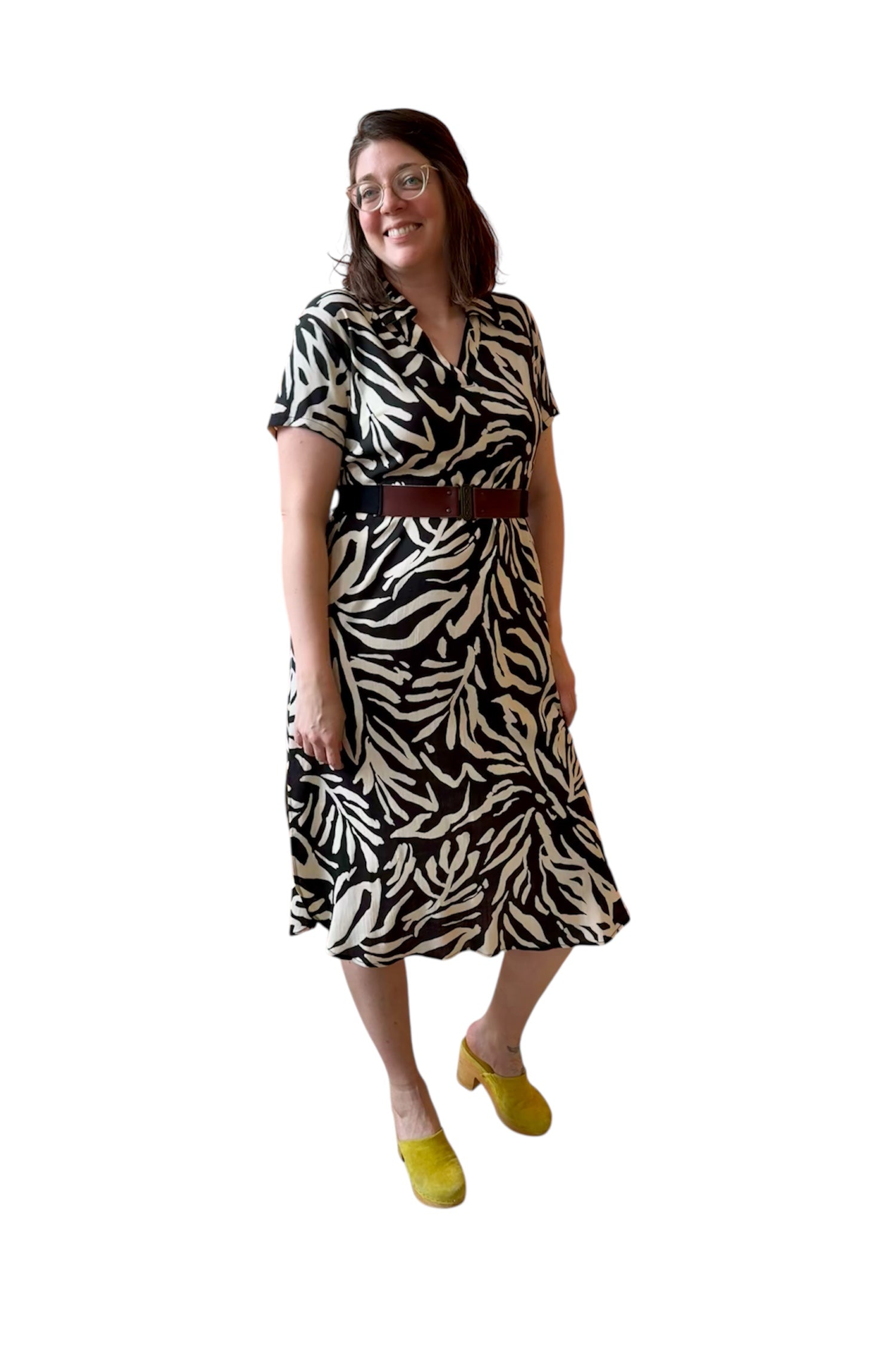 A woman wearing the Regina Dress by Compli K in Black and White print, a long dress with a v placket neck, short sleeves, loose fit, rounded hem and side slits. She is wearing it with a brown belt and standing against a white background. 