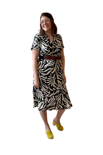 A woman wearing the Regina Dress by Compli K in Black and White print, a long dress with a v placket neck, short sleeves, loose fit, rounded hem and side slits. She is wearing it with a brown belt and standing against a white background. 