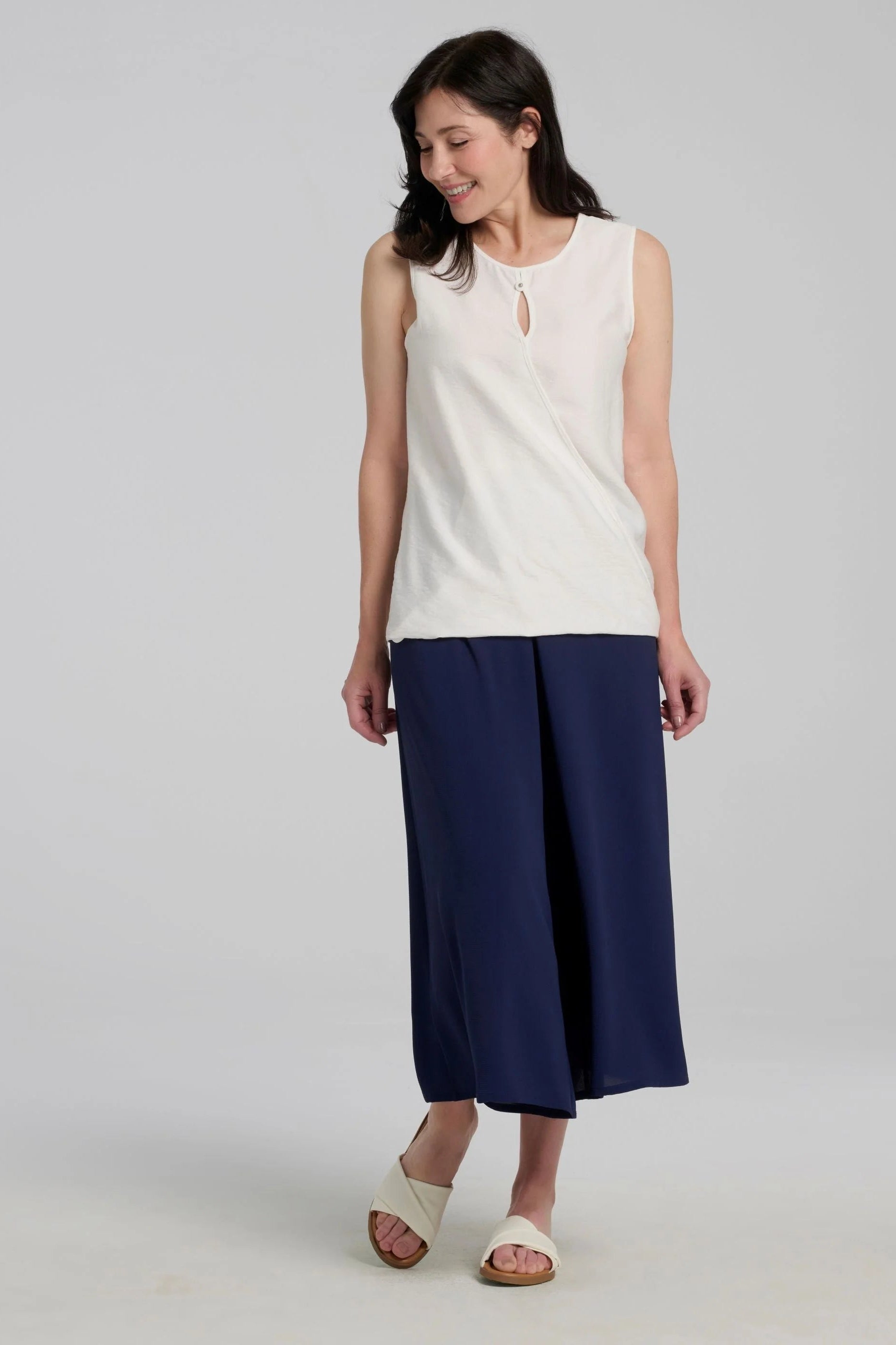 Woman wearing the Ishara Pants by Kollontai, cropped palazzo pants with ultra-wide legs and an elastic waistband. She's wearing them with a white sleeveless top and standing in front of a grey background. 
