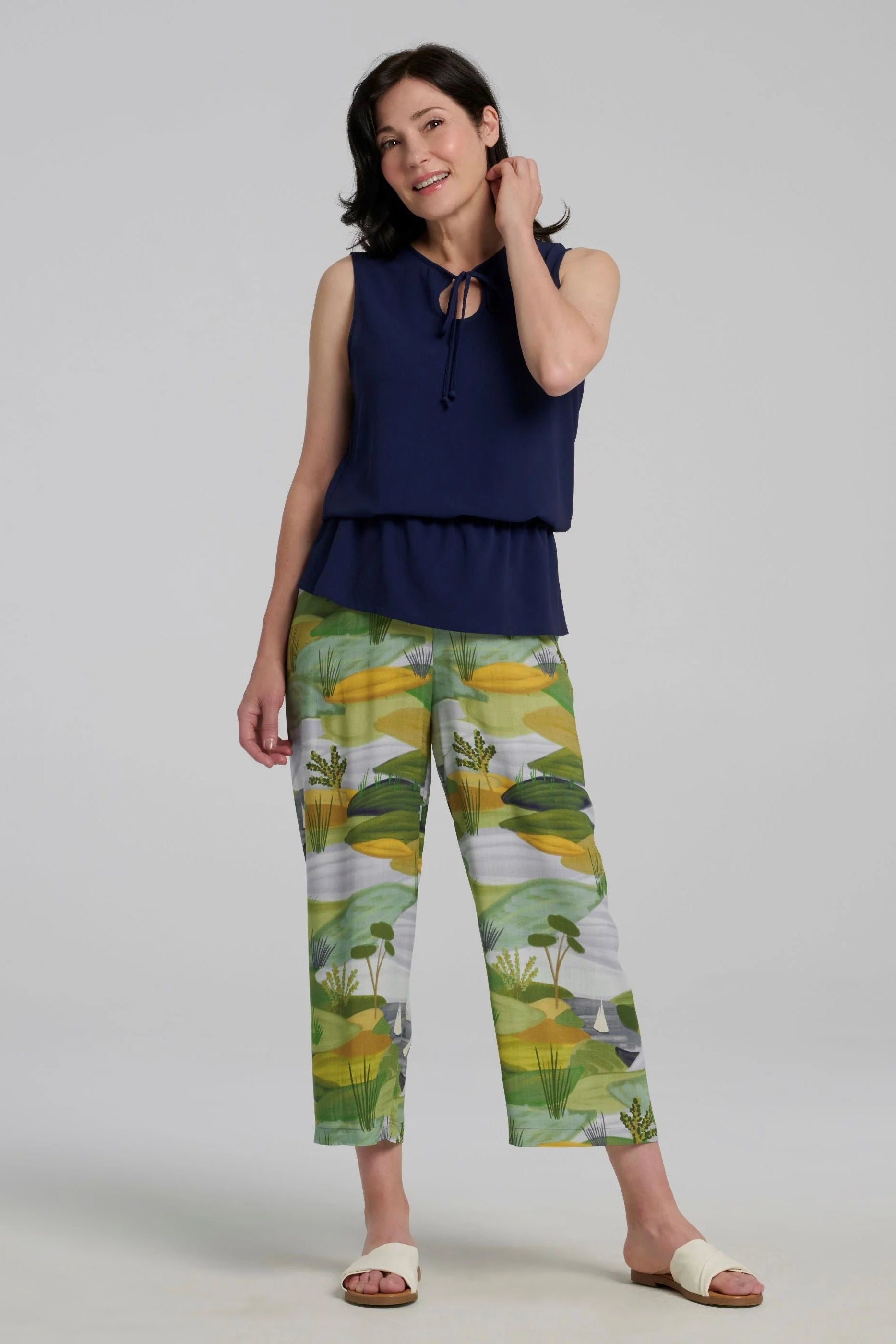 A woman wearing the Serena Top from Kollontai in Navy, a sleeveless top with a keyhole and tie neckline, elastic waist and flared peplum hem. She is wearing it with the Paraiso pants and standing against a grey background