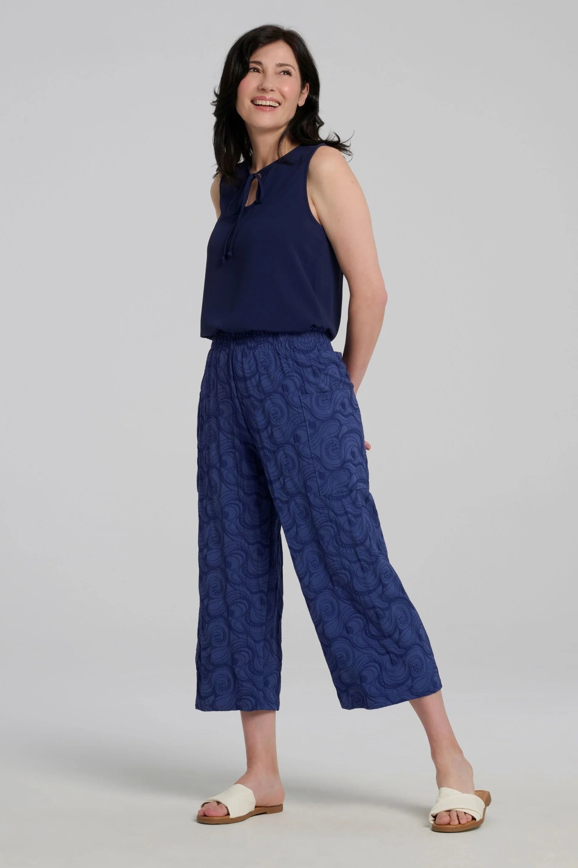 A woman wearing the Serena Top from Kollontai in Navy, a sleeveless top with a keyhole and tie neckline, elastic waist and flared peplum hem. She is wearing it with the Sana pants and standing against a a grey background