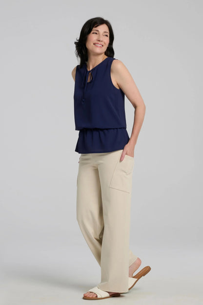 A woman wearing the Serena Top from Kollontai in Navy, a sleeveless top with a keyhole and tie neckline, elastic waist and flared peplum hem. She is wearing it with beige pants and standing against a a grey background