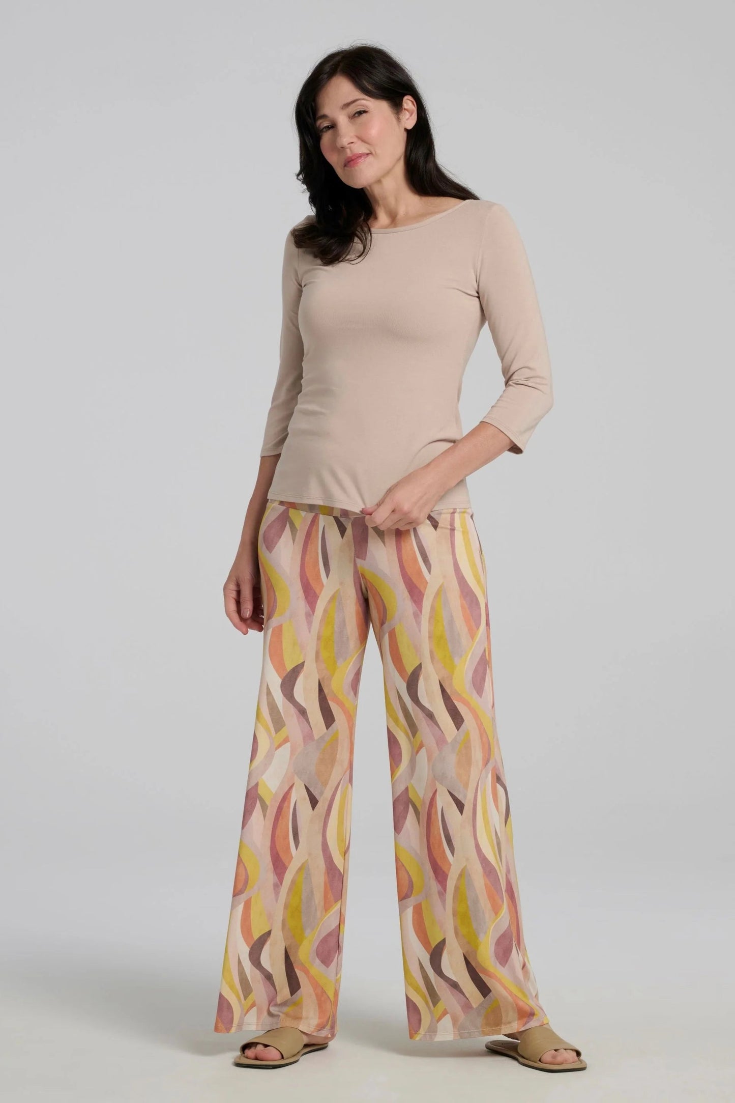 A woman wearing the Sorela pants from Kollontain in Peach print, with a wide pull-on waistband, flowing legs, and swirling geometric print. She is wearing them with a beige top and standing in front of a white background. 