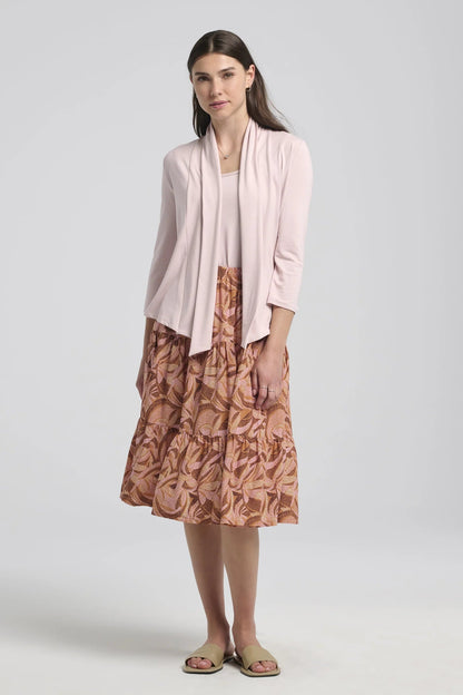 A woman wearing the Al-Uzza Skirt by Kollontai in Cinnamon print, a tiered skirt with an elastic waist and a below-the-knee length, with a pink top, standing in front of a white background 