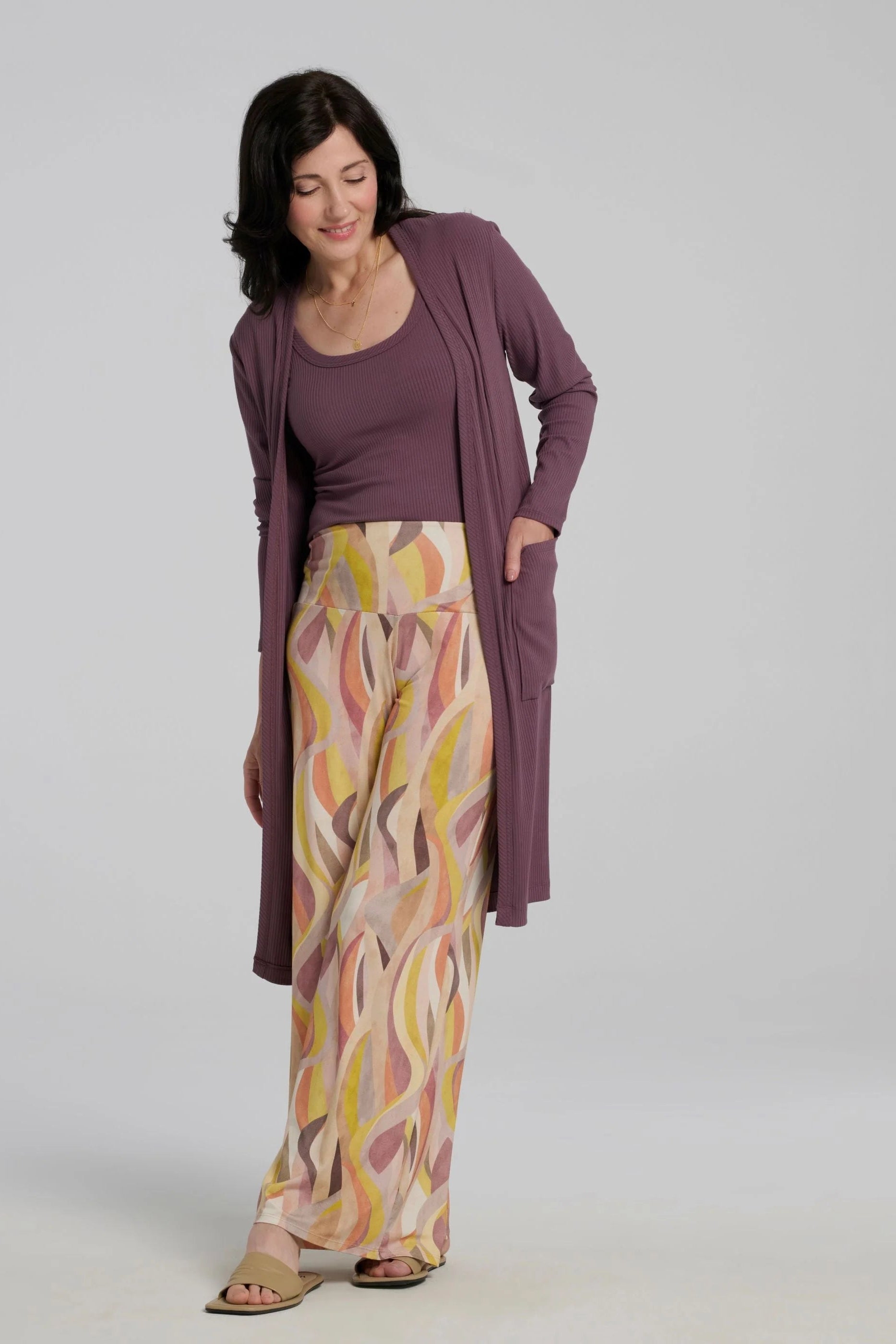 A woman wearing the Rina Camisole from Kollontai, a ribbed tank in stretchy bamboo with side straps and a scooped neck front and back.  She is wearing it with the Sorela Pants and a matching cardigan and standing in front of a white background. 