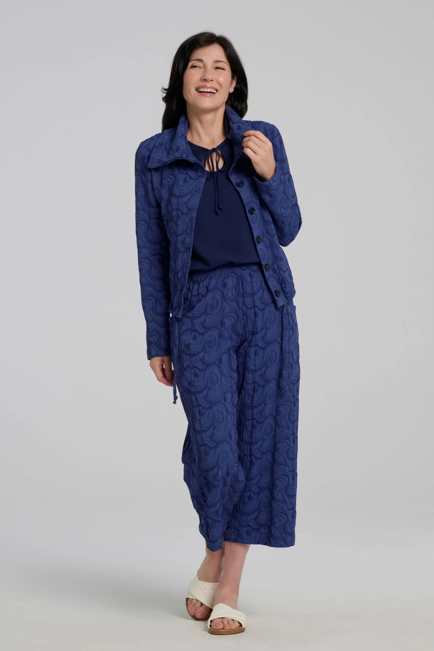 Woman wearing the Sana Pants by Kollonta in Atlantis Blue, slightly cropped straight leg pants with side patch pockets in a curlicue patterned jacquard fabric. She is wearing them with a matching jacket and standing in front of a grey background.