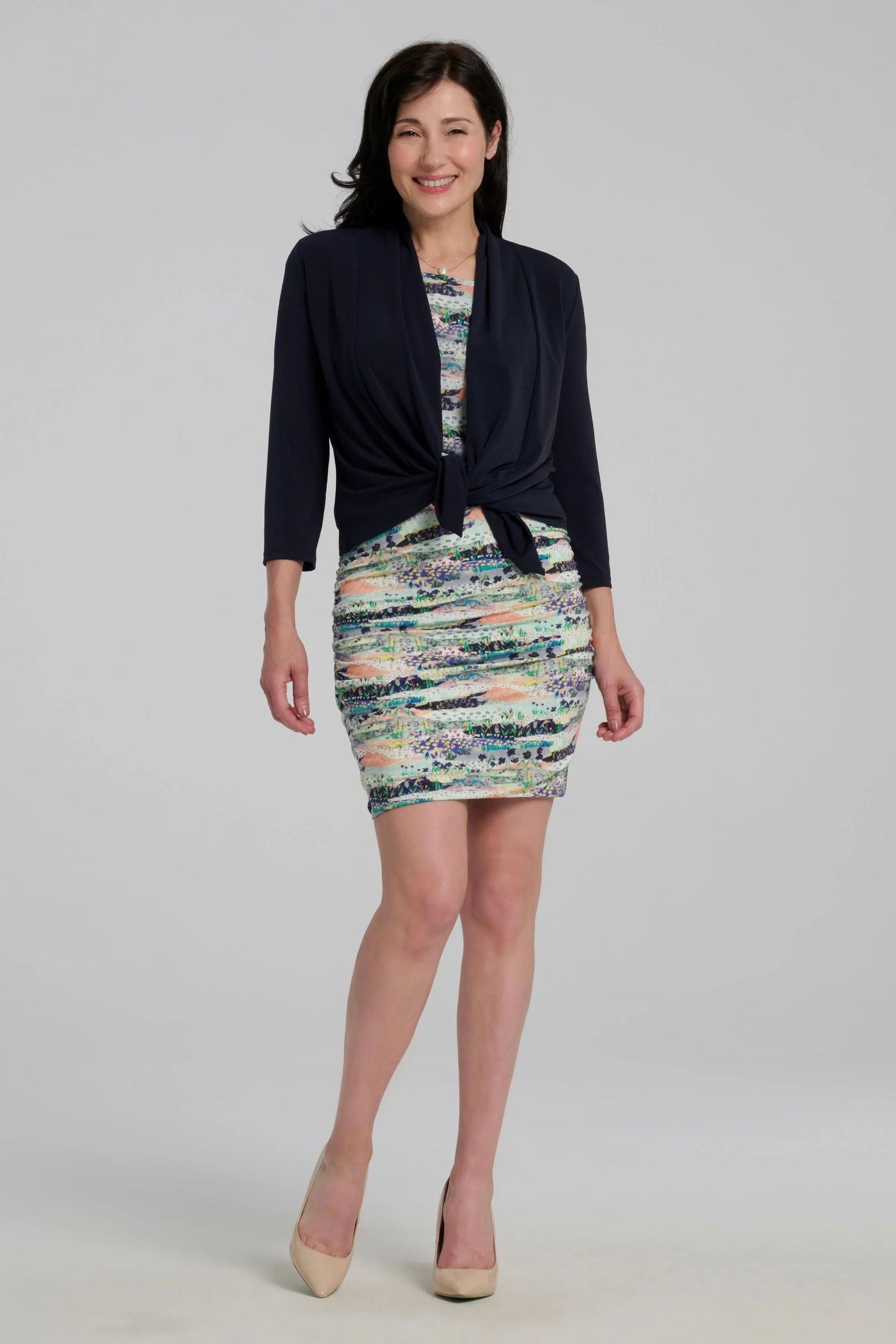 A woman wearing the Lioren Cardigan by Kollontai in Navy, a stretchy cropped jacket with longer pointed panels in the front that can be worn loose or knotted. She is wearing it over a printed dress and standing in front of a grey background 