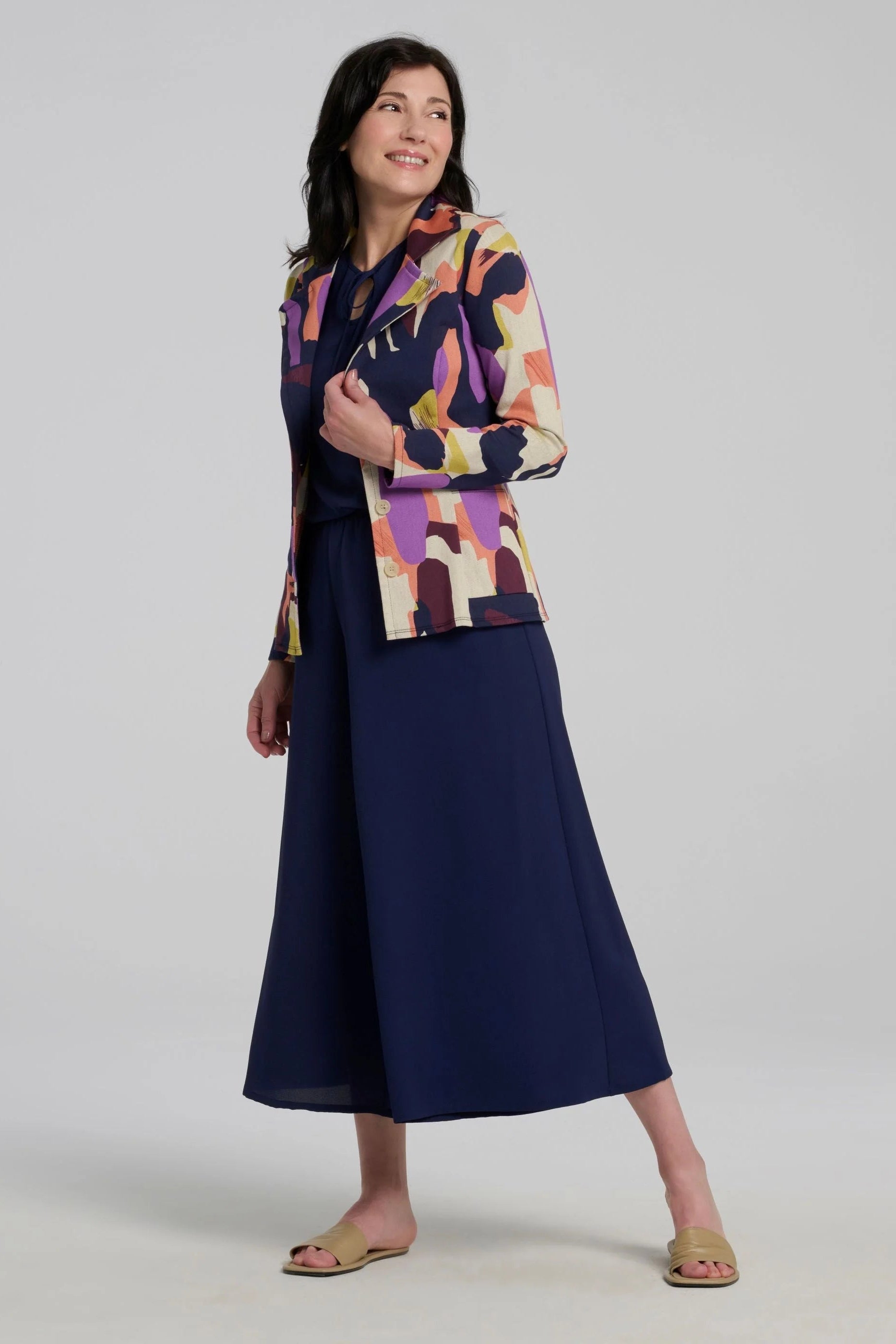 Woman wearing the Ishara Pants by Kollontai, cropped palazzo pants with ultra-wide legs and an elastic waistband. She's wearing them with a colourful blazer and standing in front of a grey background. 