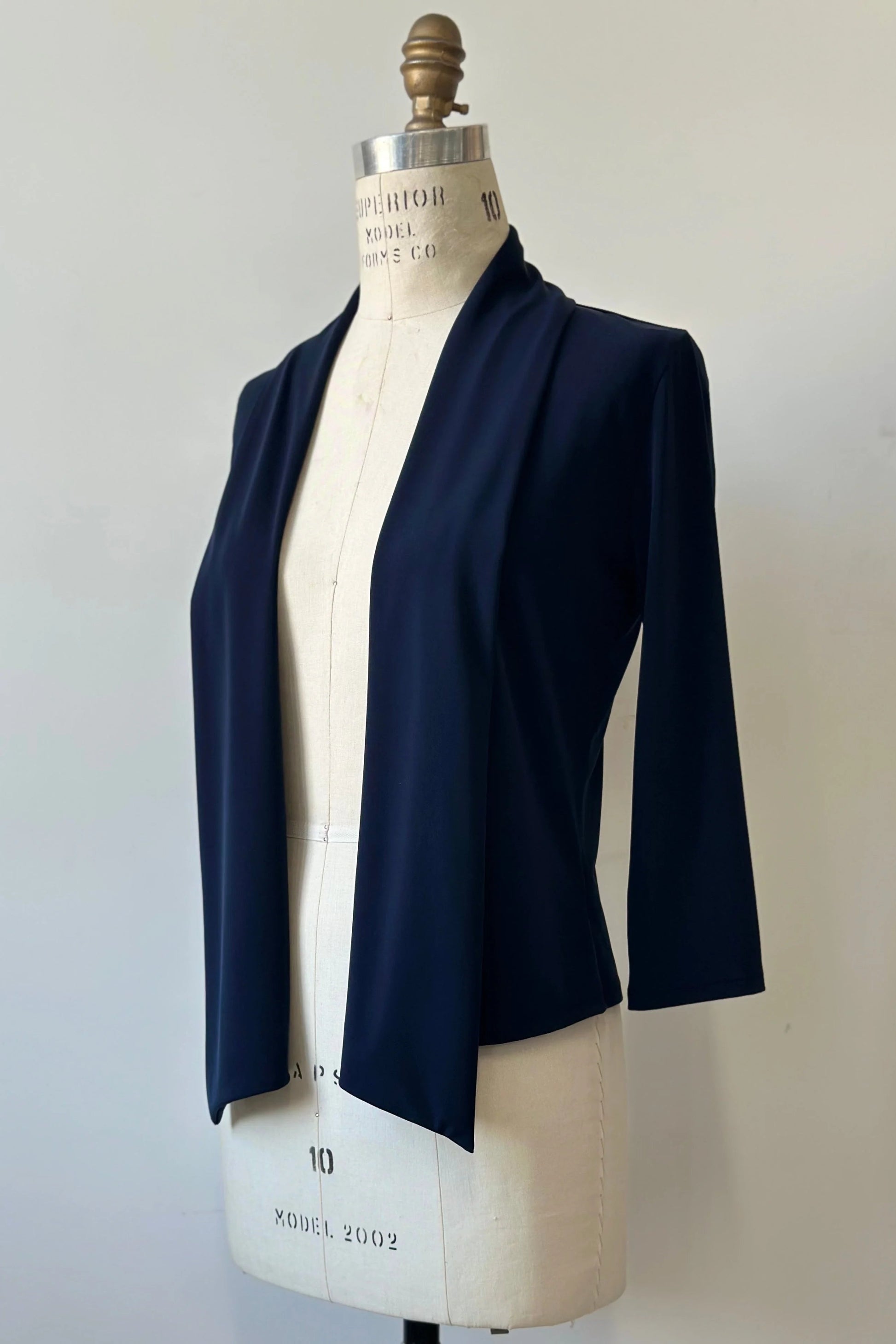 The Lioren Cardigan by Kollontai in Navy, a stretchy cropped jacket with longer pointed panels in the front that can be worn loose or knotted, is shown on a mannequin in front of a white background. 