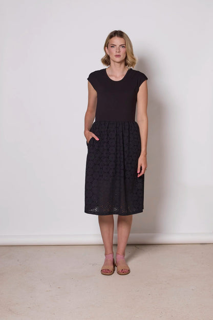 A woman wearing the Primrose Dress by Tangente in Black, a below the knee length dress featuring a bamboo rayon bodice and daisy eyelet skirt, with short sleeves and a round neck, standing against a plain background. 