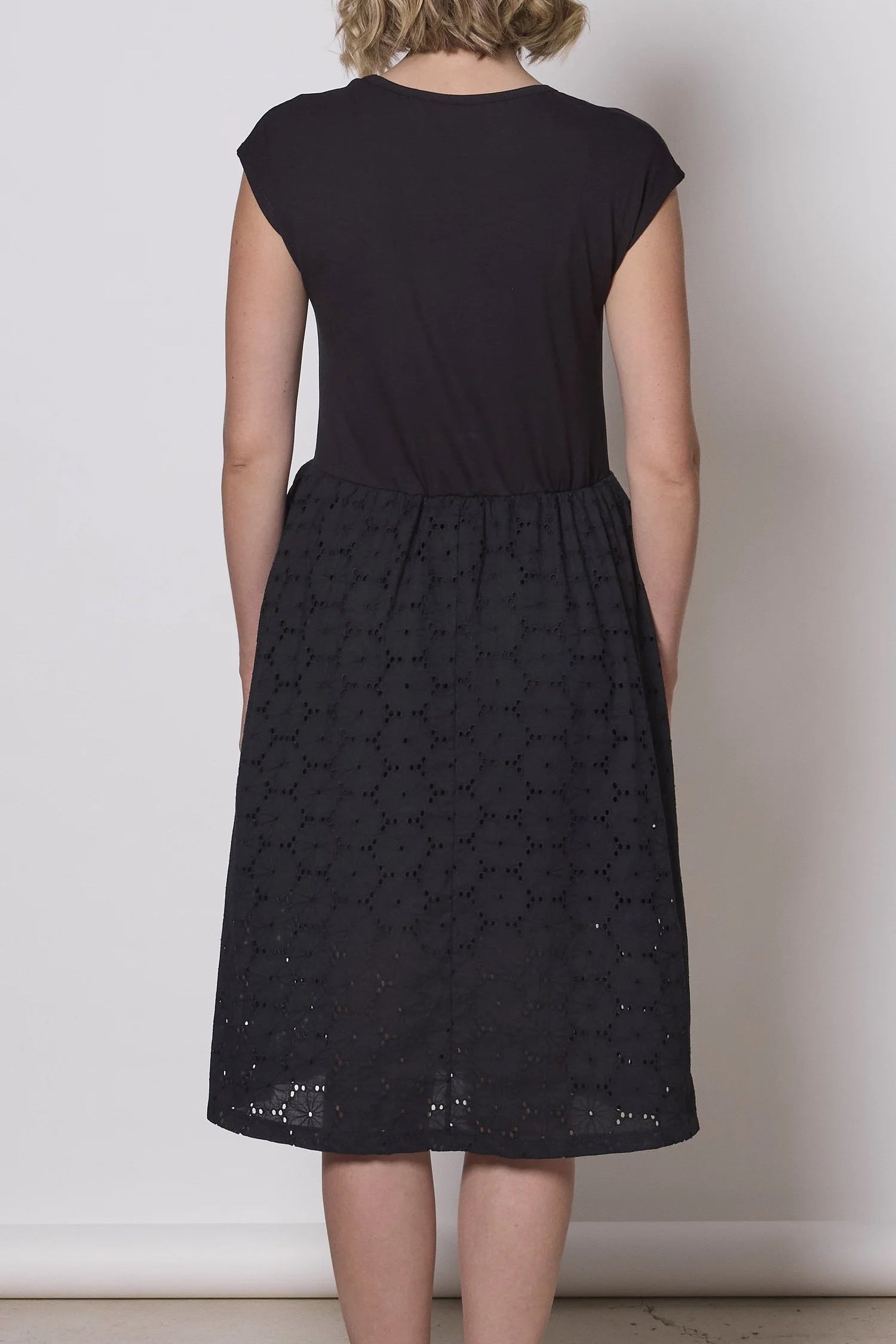 Back view of a woman wearing the Primrose Dress by Tangente in Black, a below the knee length dress featuring a bamboo rayon bodice and daisy eyelet skirt, with short sleeves and a round neck, standing against a plain background. 