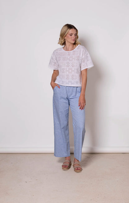 A woman wearing the Florence Top by Tangente in White, an embroidered eyelet top with short drop sleeves and a boxy fit. She is wearing it with light blue pants against a plain background. 