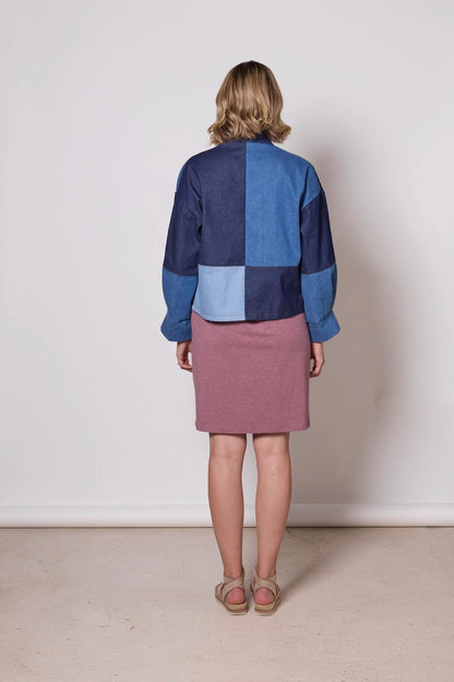 Back view of a woman wearing the Myrtle Denim Jacket by Tangente, a patchwork jacket with an open front, drop sleeves, and front patch pockets. She is wearing it over a pink against a white background