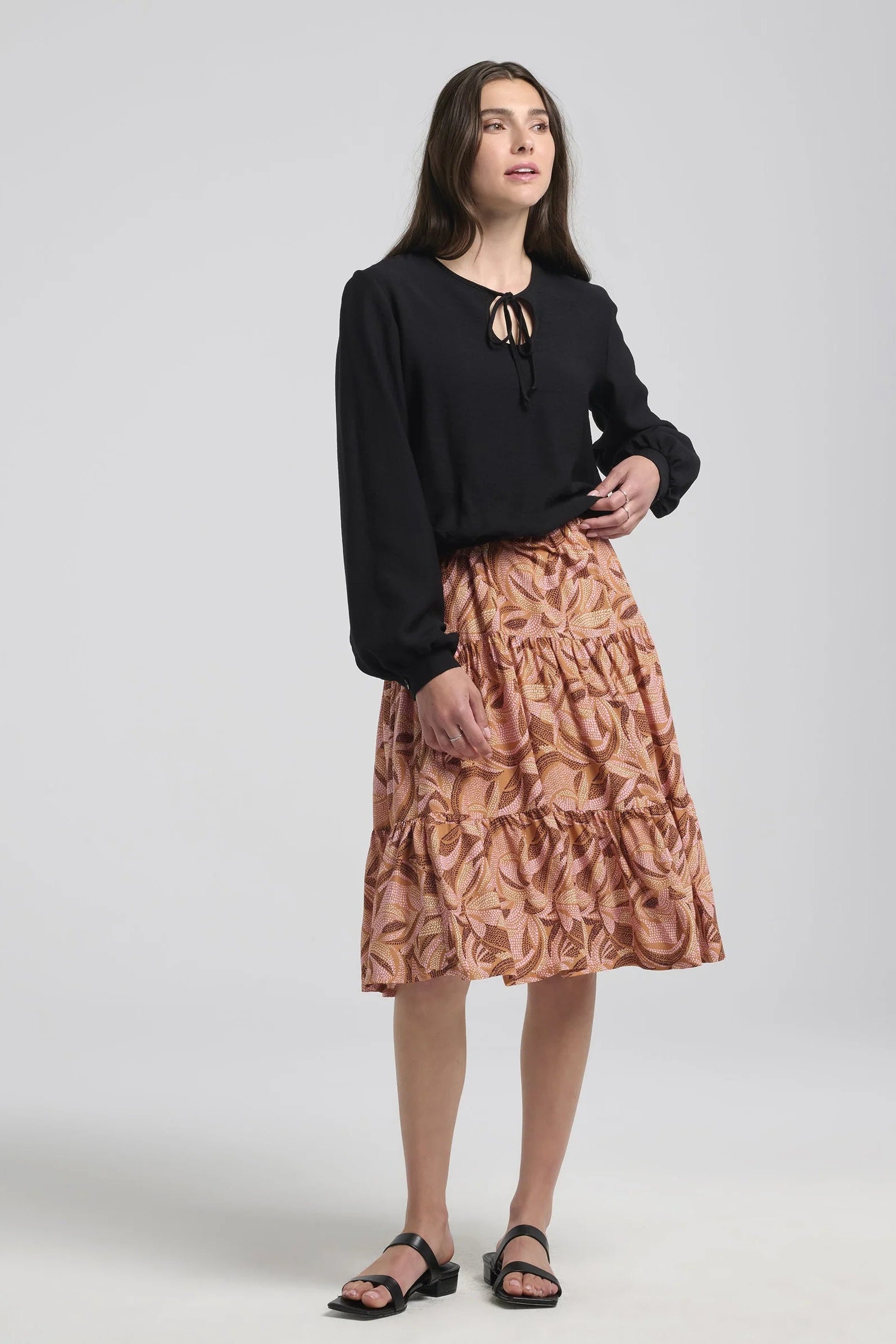 A woman wearing the Al-Uzza Skirt by Kollontai in Cinnamon print, a tiered skirt with an elastic waist and a below-the-knee length, with a black top, standing in front of a white background 