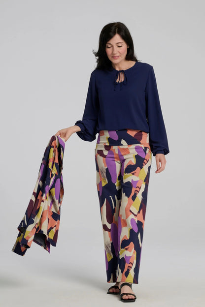 A woman wearing the Elma Pants by Kollontai in a Multicolour abstract print, with a wide pull-one waistband, straight legs, and slanted pockets. She is wearing them with a navy top and 
and holding a matching jacket while standing in front of a plain background